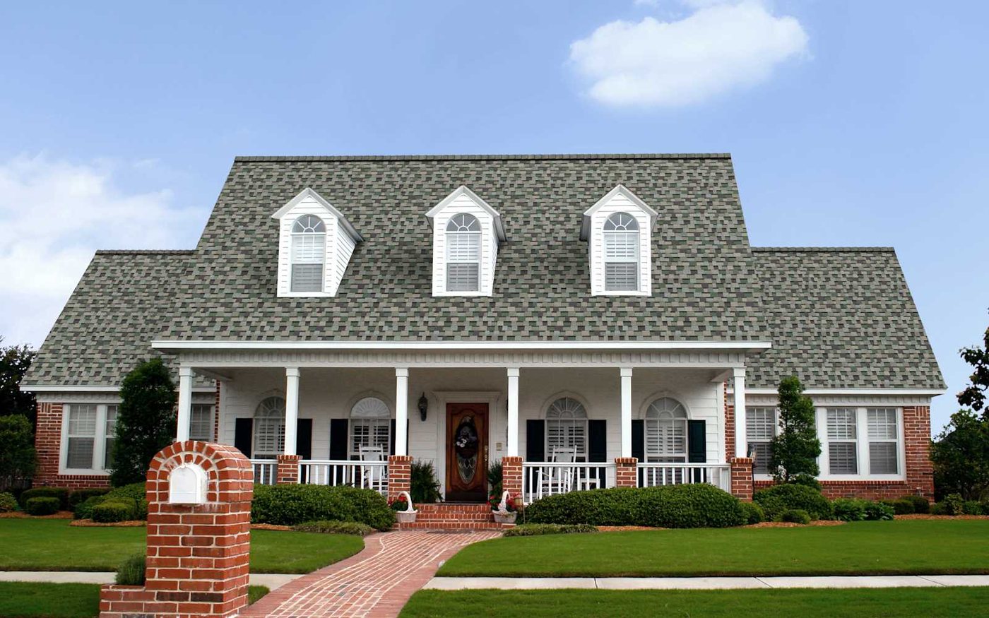 A white farmhouse with red brick accents roofed in Owens Corning TruDefinition Duration Designer shingles in Evergreen Mist